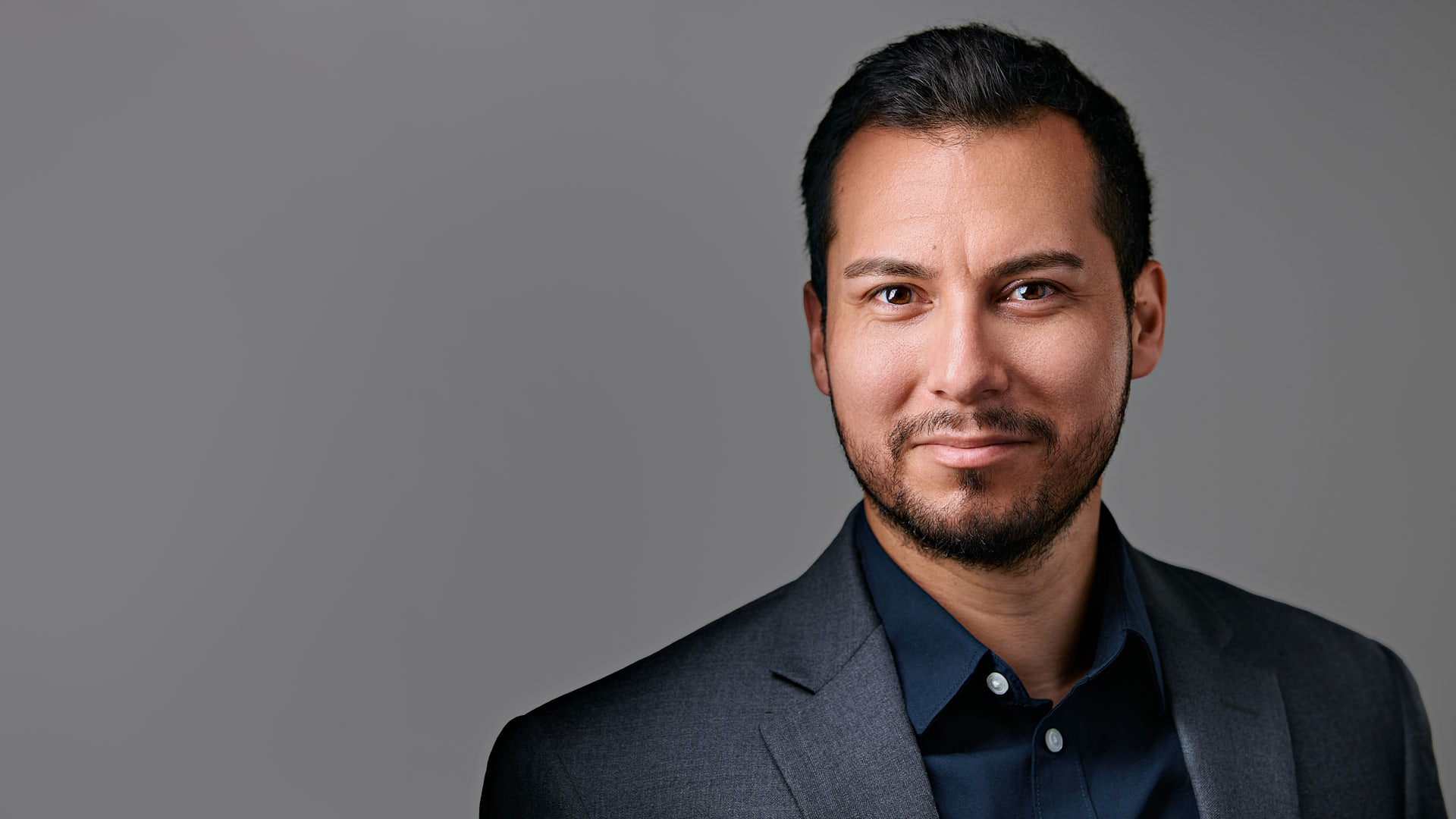 Professional headshot of a man in a dark gray suit. Professional headshot of a man with short dark hair and a trimmed beard, wearing a navy shirt and gray blazer, smiling confidently in a studio setting in Stanford.