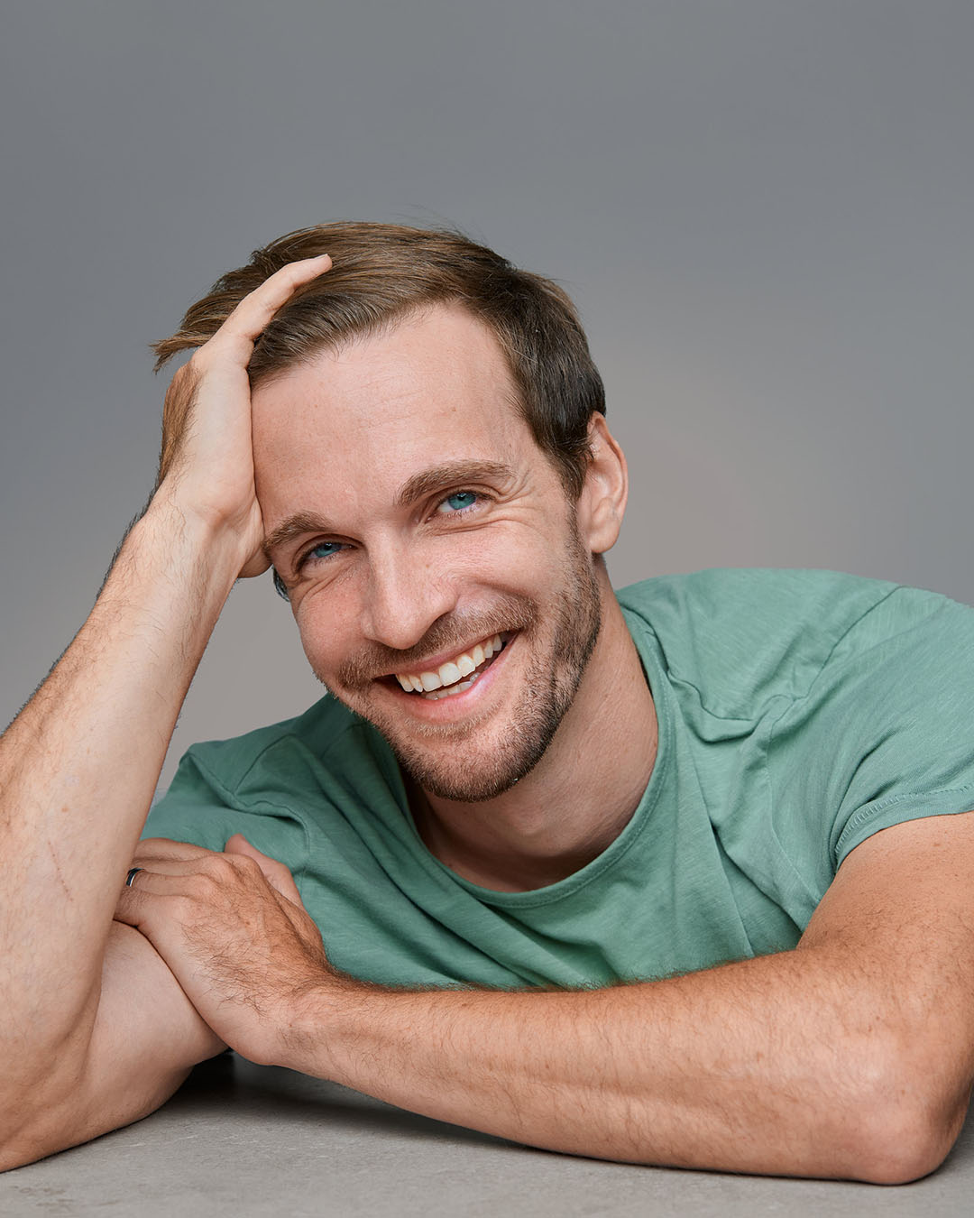 Professional Headshot by Dean Birinyi - Palo Alto, Silicon Valley A smiling man in a green t-shirt, resting his head on one hand and leaning on a surface, captured in a professional headshot by Dean Birinyi. Neutral gray background emphasizes his approachable and confident expression.