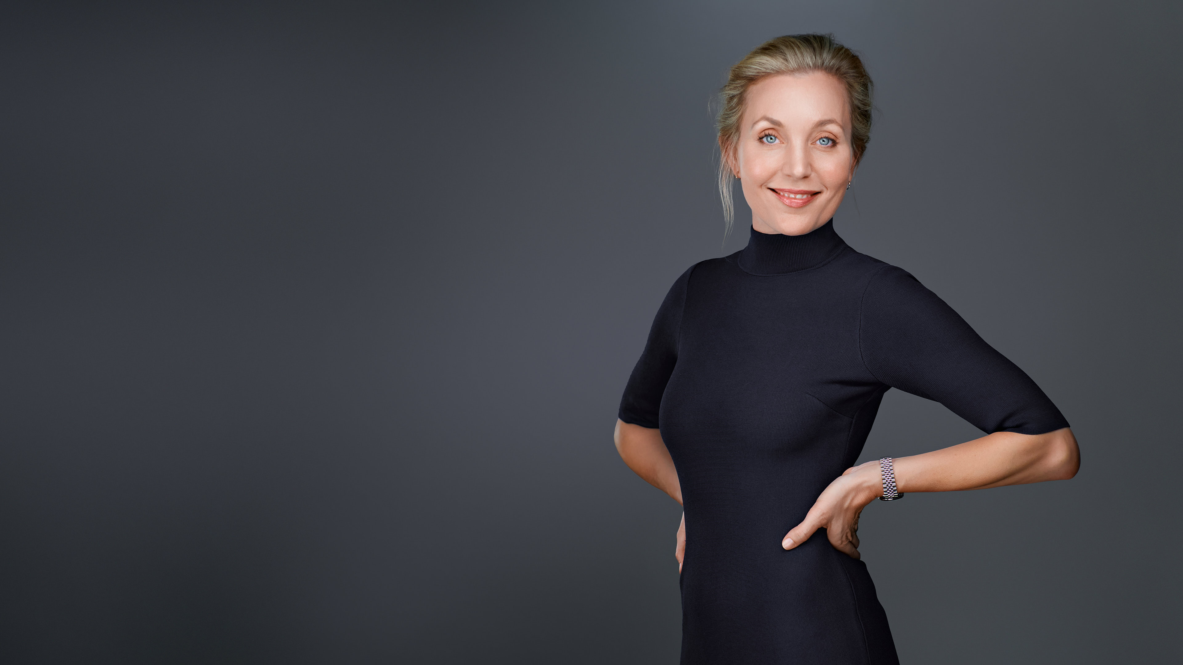 Professional Headshot by Dean Birinyi - Palo Alto, Silicon Valley Professional headshot of a woman with blonde hair tied back, wearing a black fitted dress and a silver watch, standing confidently against a gray studio backdrop in Stanford.