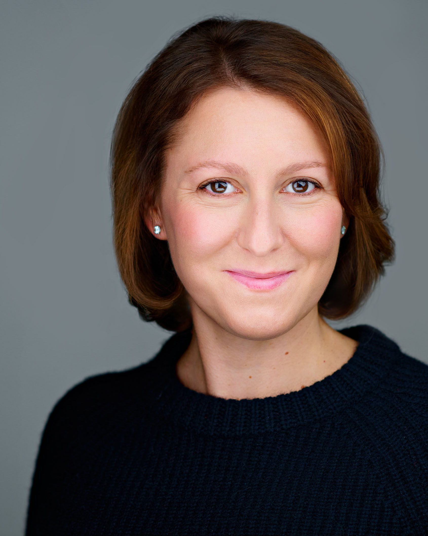 Professional Headshot by Dean Birinyi - Palo Alto, Silicon Valley A professional woman with blonde hair, styled neatly, wearing a fitted black dress with a high neckline. She stands confidently with her hands on her hips, against a neutral gray background.