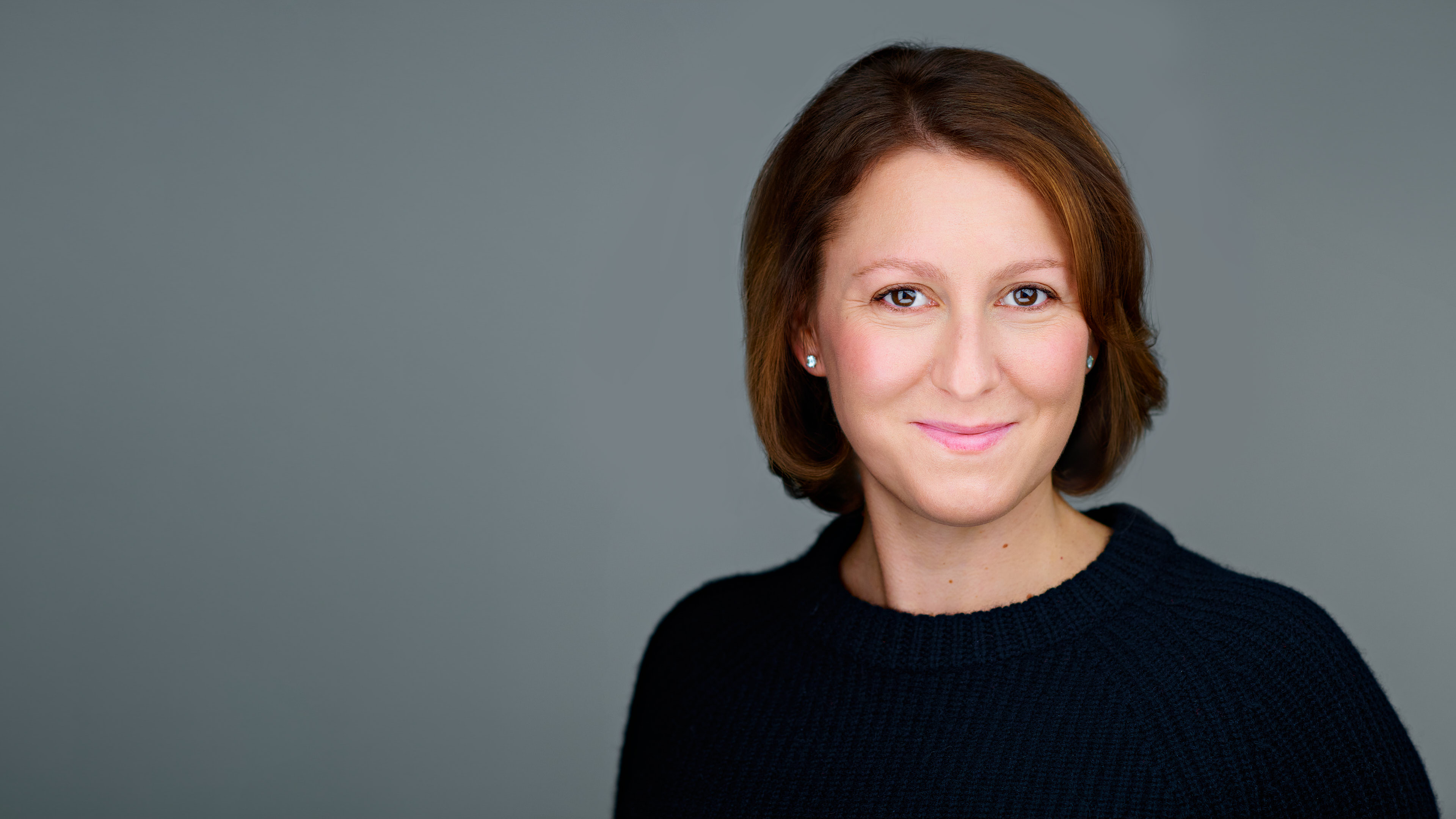 Professional Headshot by Dean Birinyi - Palo Alto, Silicon Valley Professional headshot of a woman with light brown hair, wearing a navy sweater, smiling warmly against a gray background in Stanford, photographed by Dean Birinyi Photography