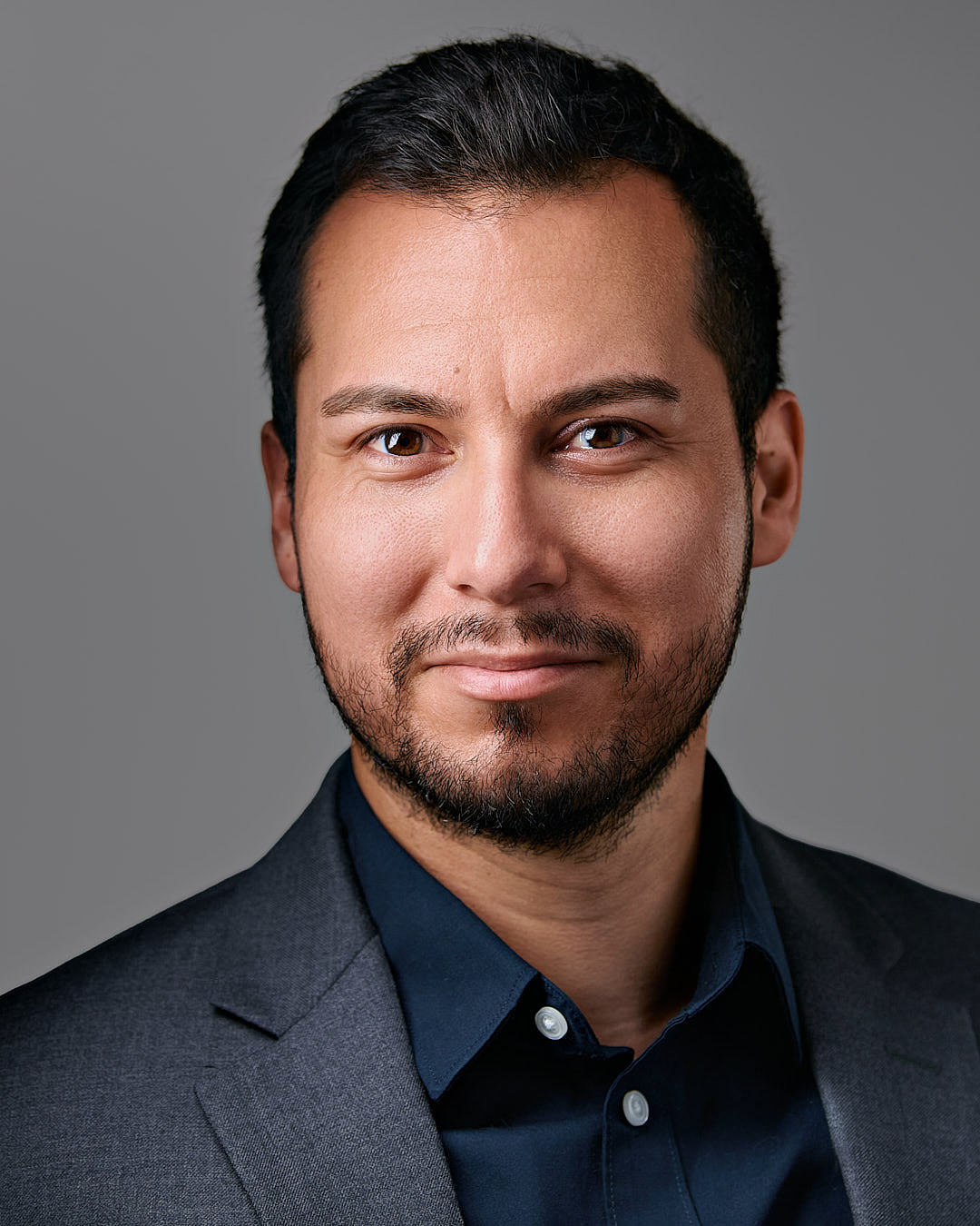 Professional headshot of a man in a dark gray suit. A man with short dark hair and a trimmed beard, wearing a dark gray suit jacket and a navy blue button-up shirt, smiles subtly while looking directly at the camera against a gray background.