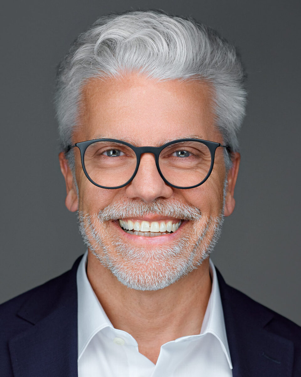 A middle-aged man with gray hair and a beard wearing glasses, smiling broadly against a neutral gray background.