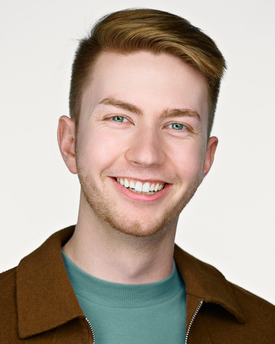 A professional headshot of a young man with light brown hair, smiling brightly while wearing a brown jacket and green shirt, against a light background
