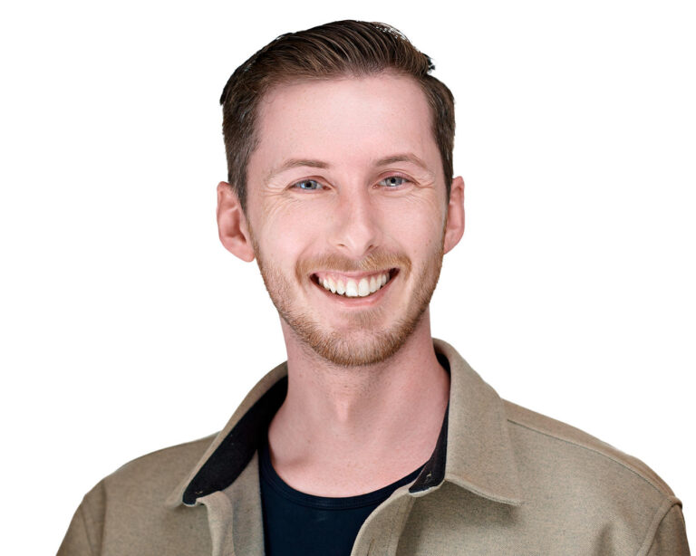 A man with short, light brown hair and a neatly trimmed beard, smiling broadly while wearing a beige jacket over a black shirt, standing against a white background.