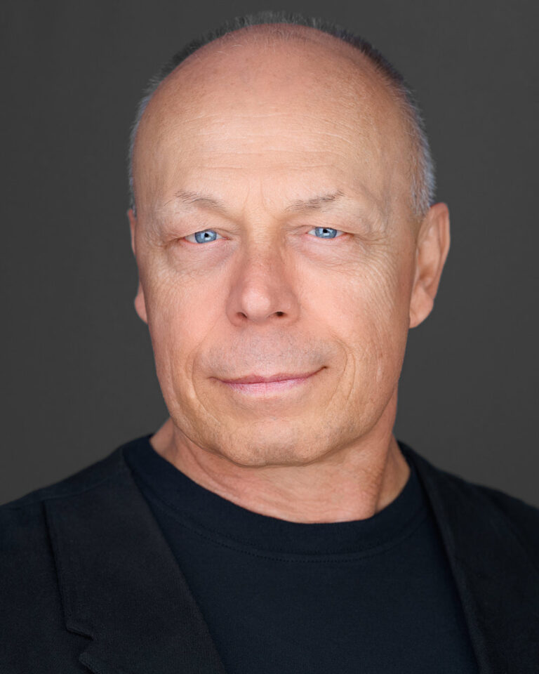 A professional headshot of Dean Birinyi, a bald man with light skin and blue eyes, wearing a black blazer over a black shirt, with a neutral facial expression, set against a dark gray background.