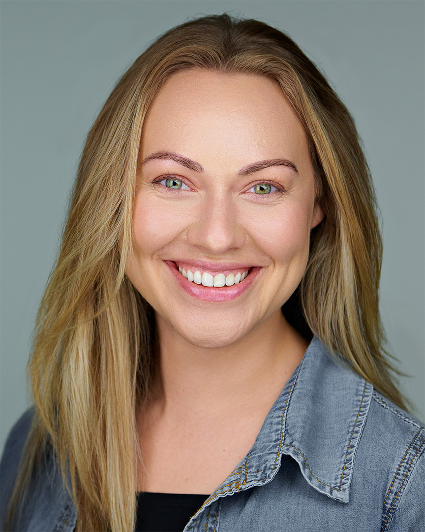 A smiling woman with blonde hair wearing a denim shirt against a soft gray background, photographed by Dean Birinyi in his Palo Alto commercial studio.