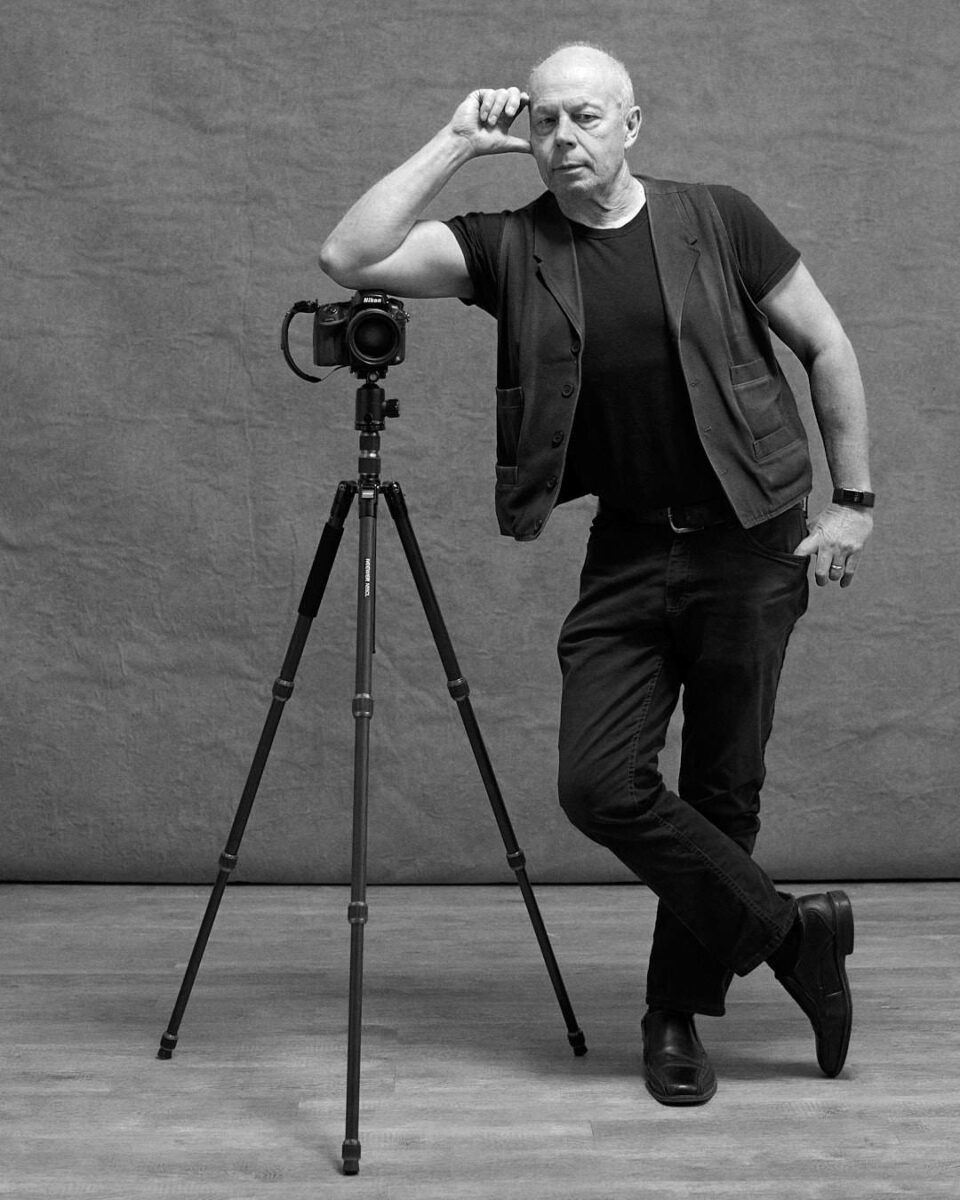 A black and white portrait of a photographer leaning casually against a tripod with a camera.