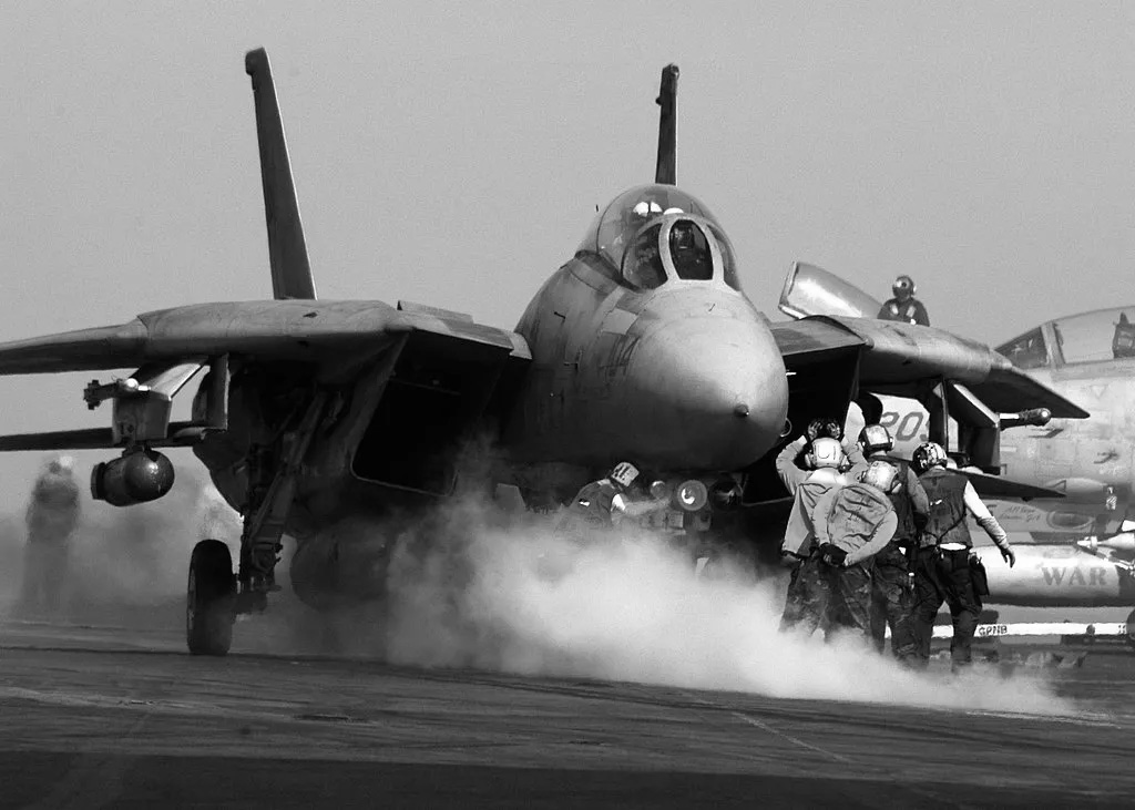 F-14 Tomcat fighter jet preparing for takeoff on an aircraft carrier deck with flight crew in position.