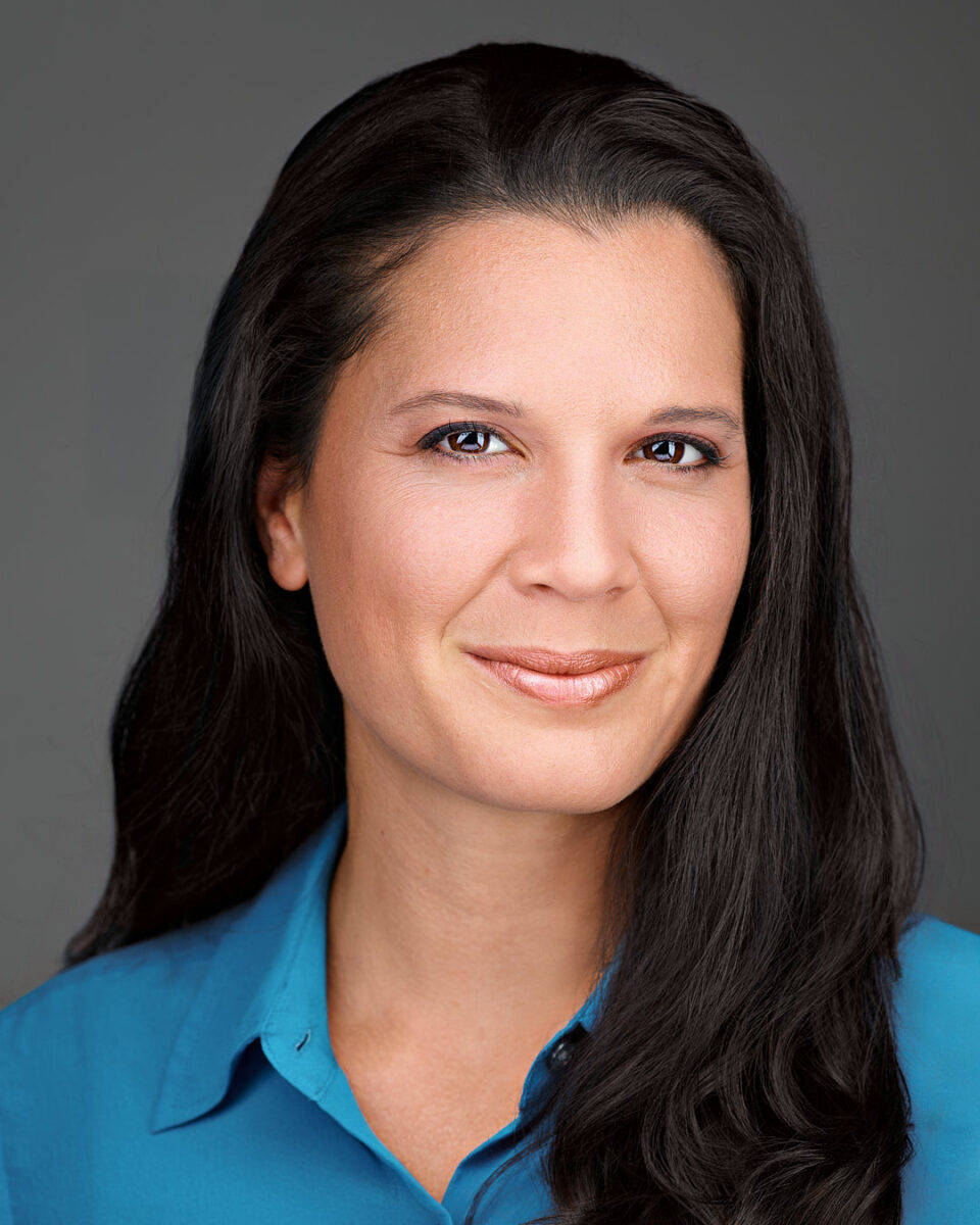 Professional headshot of a confident woman with long dark hair, wearing a blue blouse against a neutral gray background.