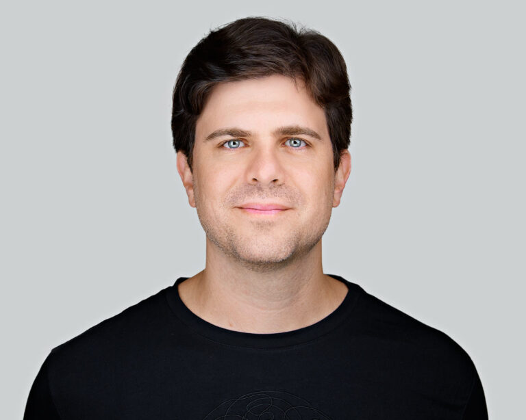 A professional headshot of a man with dark hair, wearing a black T-shirt, smiling gently against a neutral light gray background.