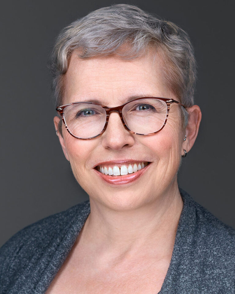 Professional headshot of a woman with short gray hair, wearing glasses and a gray sweater, smiling warmly against a neutral gray background.