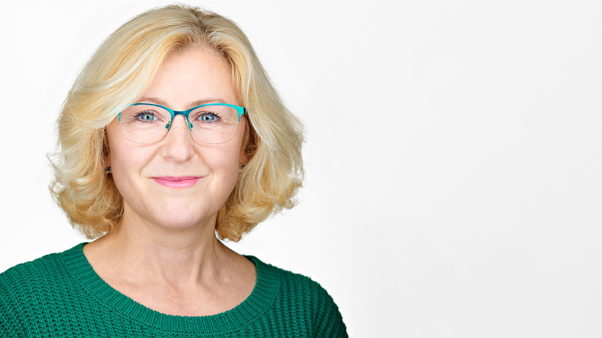 Professional headshot of a smiling woman with blonde hair and teal glasses, wearing a green sweater against a white background.