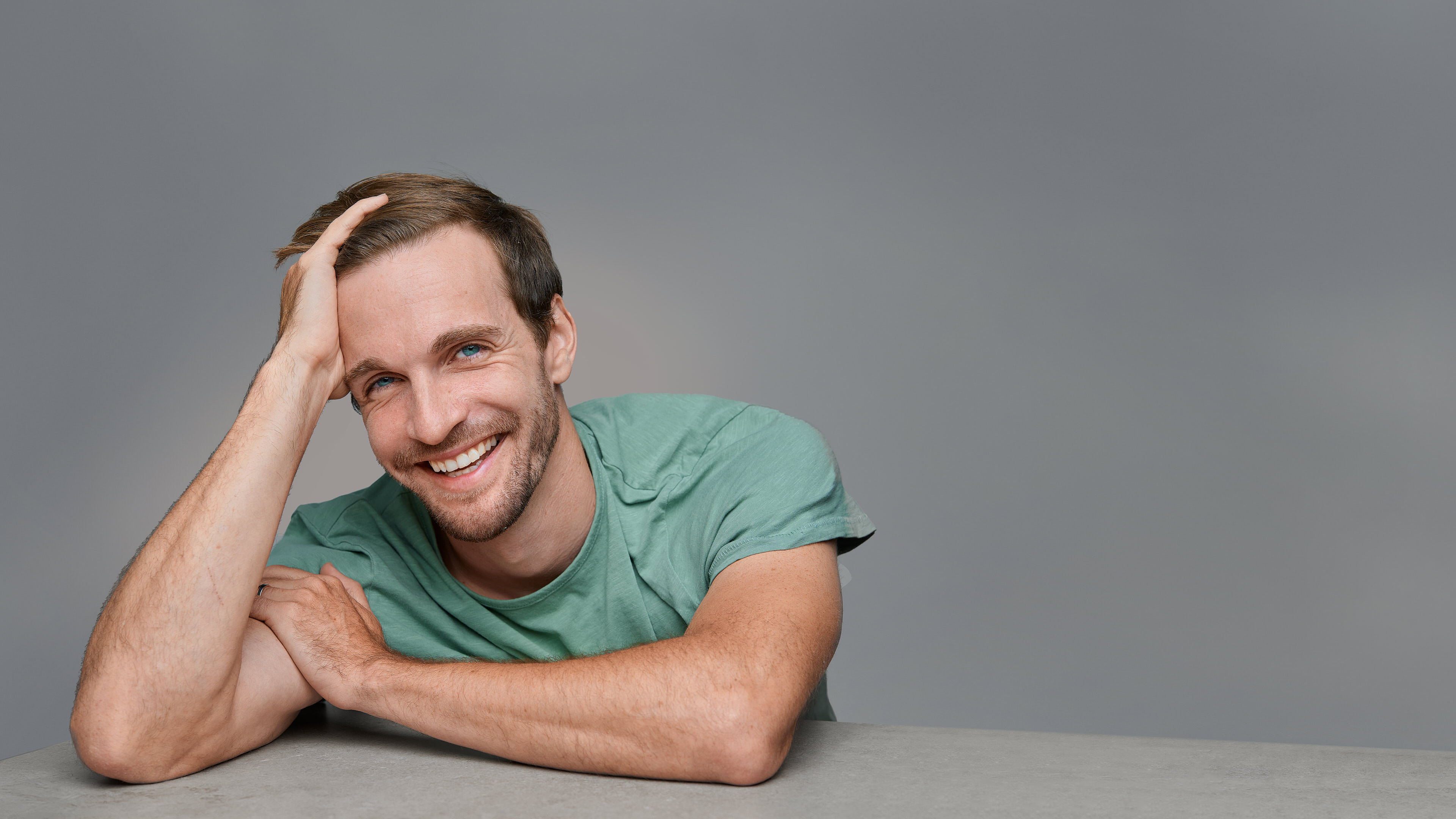 A smiling man in a green t-shirt, resting his head on one hand and leaning on a surface, captured in a professional headshot by Dean Birinyi. Neutral gray background emphasizes his approachable and confident expression.