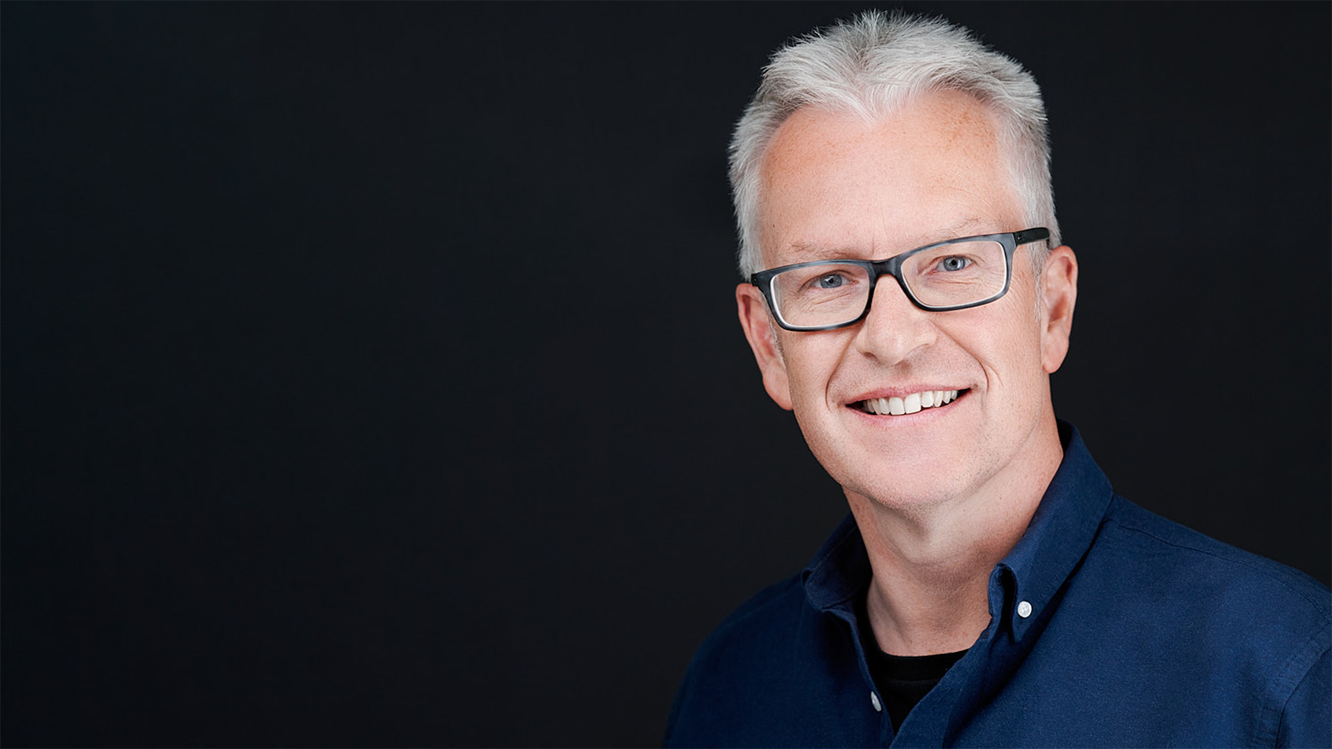 Professional headshot of a smiling man with white hair and glasses, wearing a navy blue shirt against a dark background.