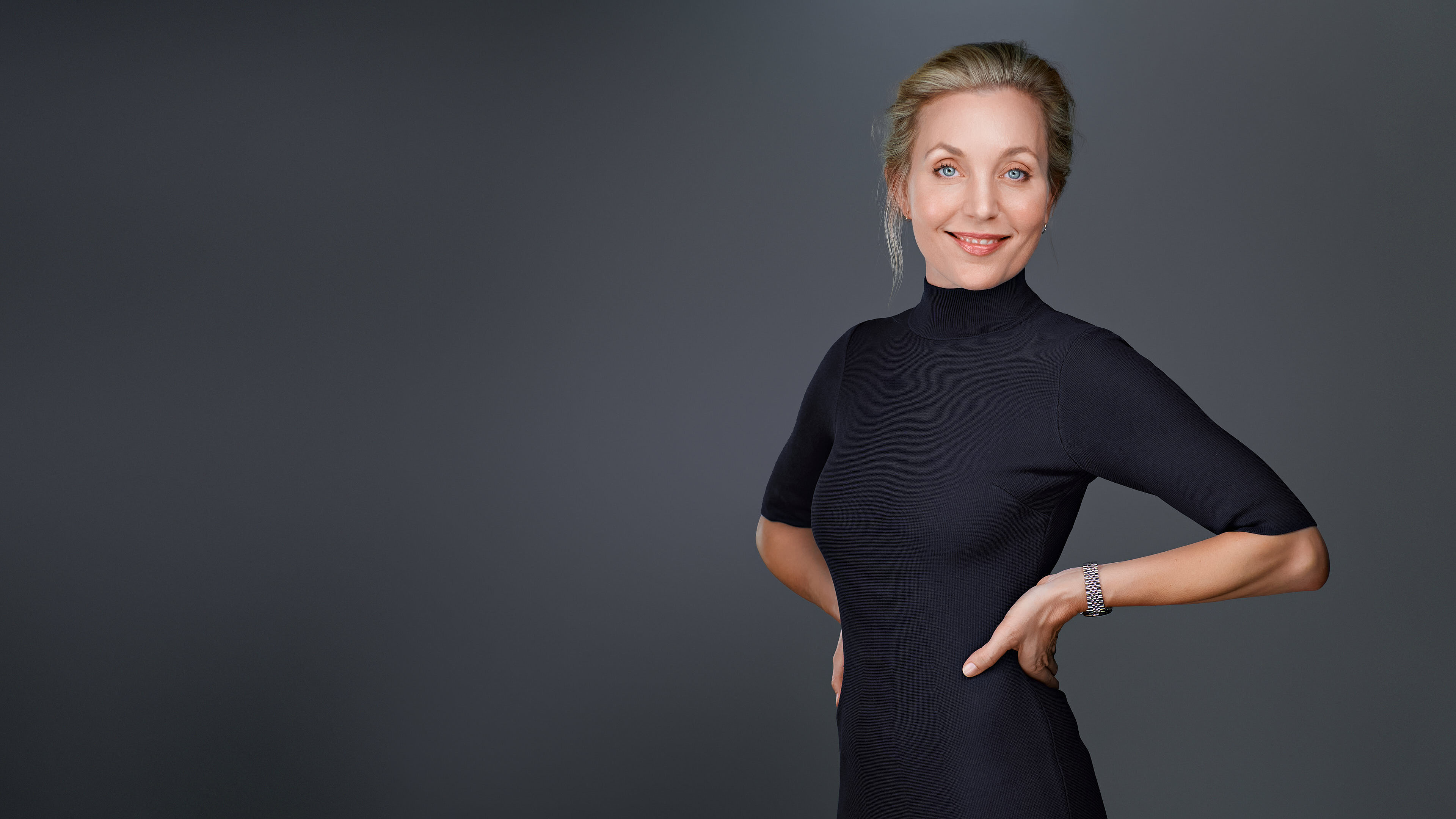 A professional woman with blonde hair, styled neatly, wearing a fitted black dress with a high neckline. She stands confidently with her hands on her hips, against a neutral gray background.