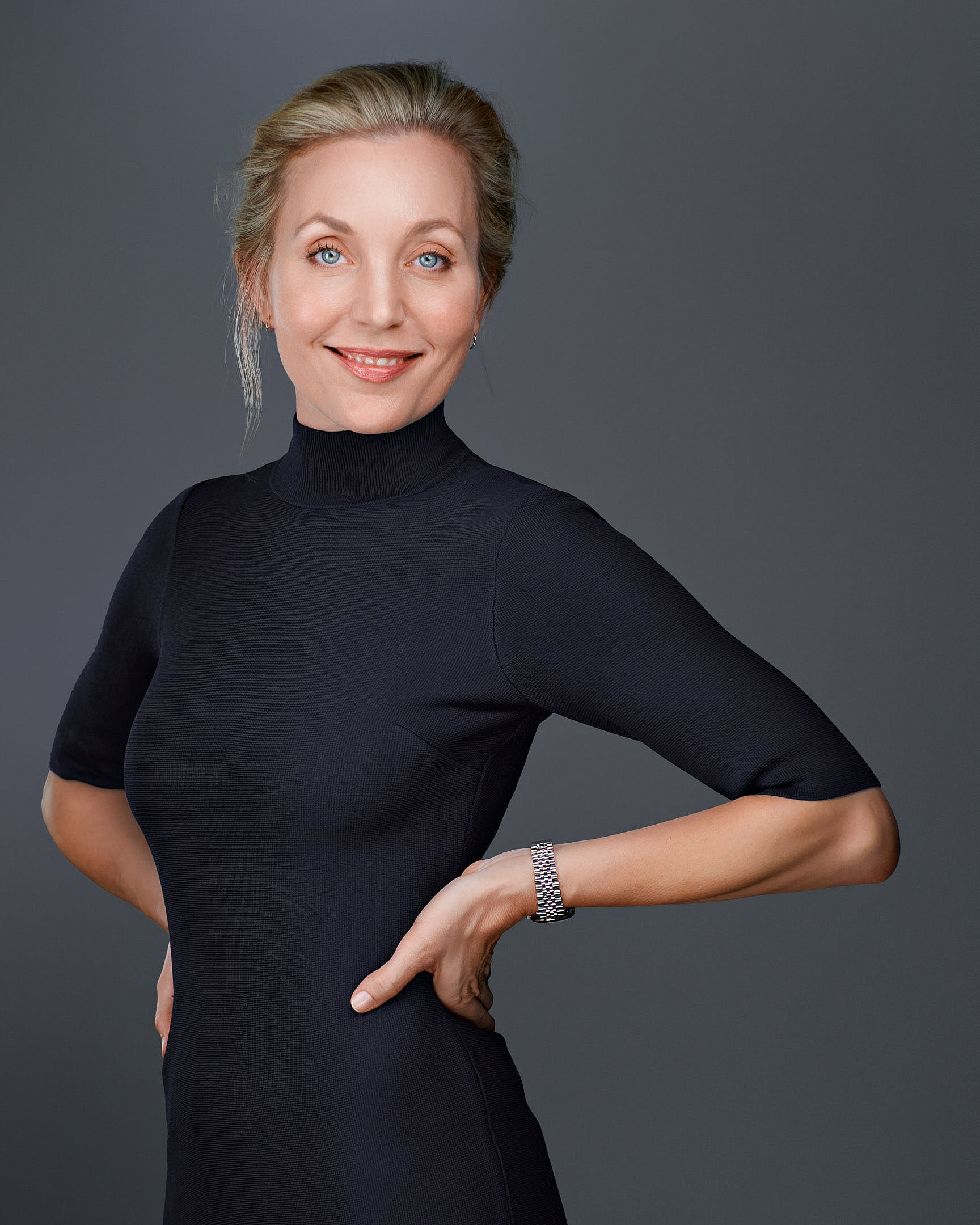 A professional woman with blonde hair, styled neatly, wearing a fitted black dress with a high neckline. She stands confidently with her hands on her hips, against a neutral gray background.