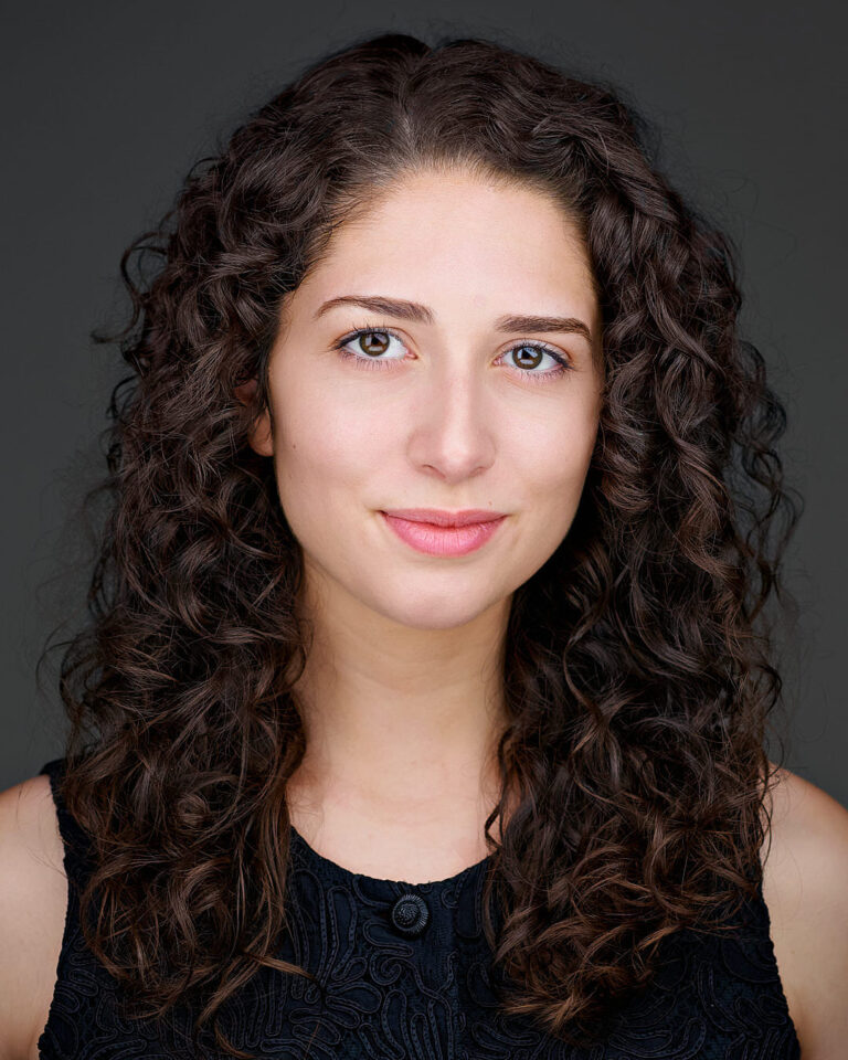 Professional headshot of a young woman with curly dark hair, wearing a black top and smiling softly against a dark background.