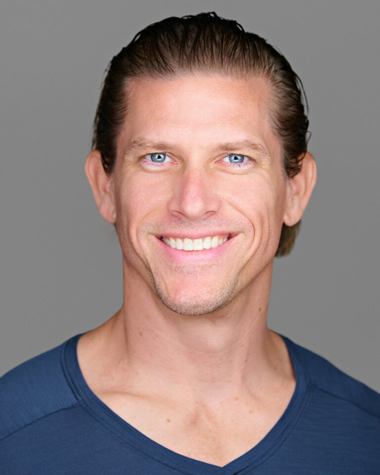 A professional headshot of a man with short hair and a bright smile, wearing a navy V-neck shirt, against a neutral gray background.