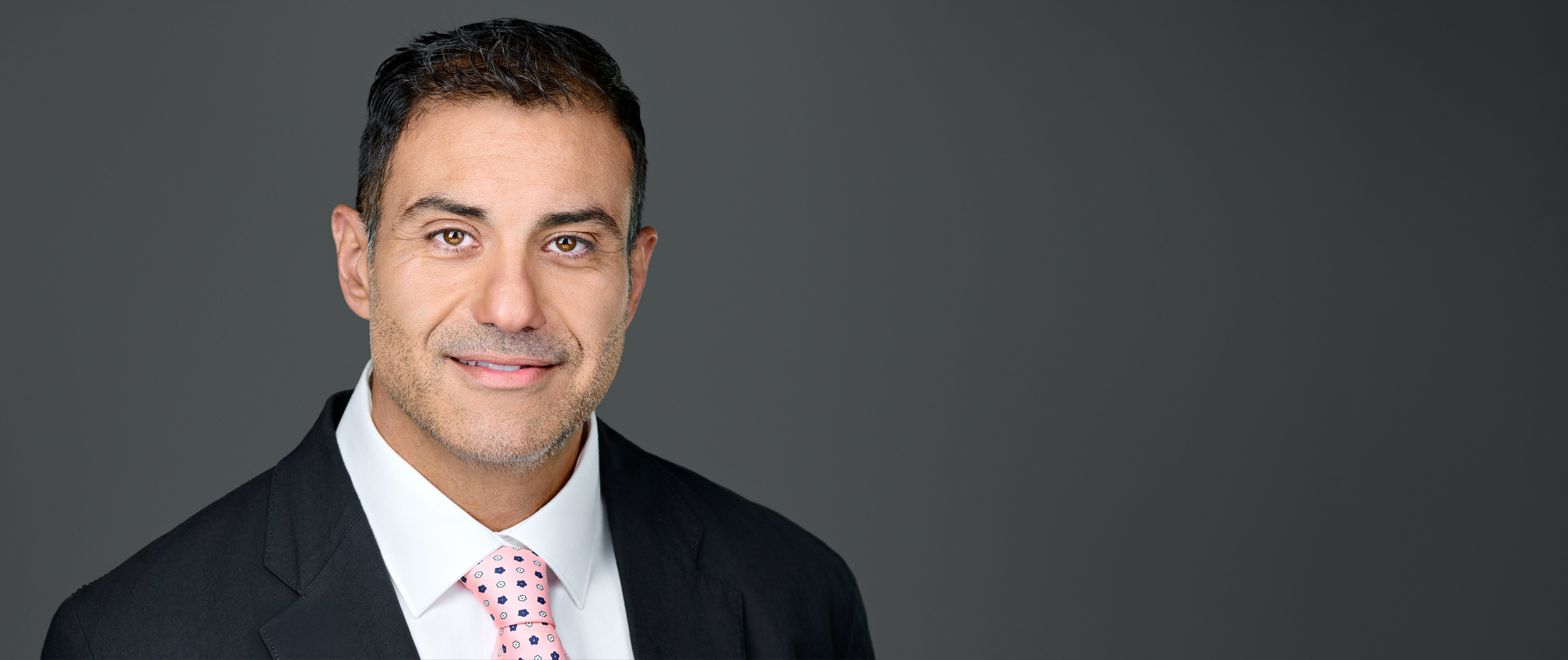 A professional headshot of a sales director with short dark hair, wearing a black suit, white shirt, and pink tie, against a neutral gray background. Photographed by Dean Birinyi in his Silicon Valley Photo Studio.