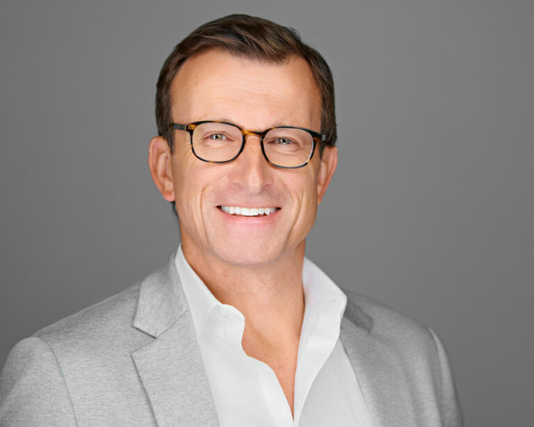Corporate headshot of a male in-house counsel in Palo Alto by Dean Birinyi, featuring a confident smile, glasses, and a light gray suit against a neutral gray background.
