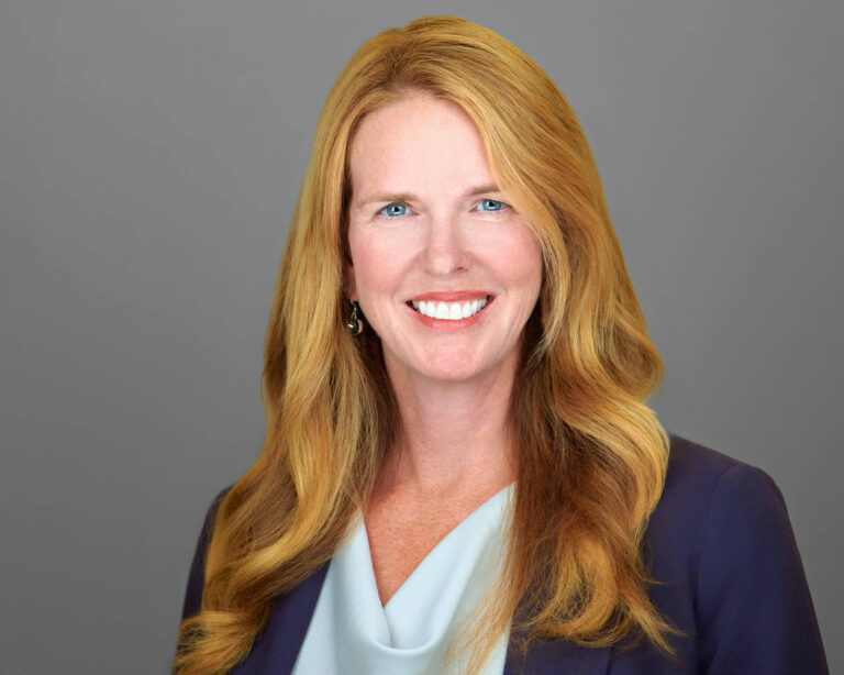 Corporate headshot of a female executive in Palo Alto by Dean Birinyi, featuring a confident smile, long red hair, and a navy blazer against a neutral gray background.