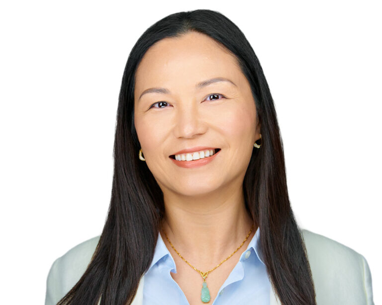 Corporate headshot of a female executive in Palo Alto by Dean Birinyi, featuring a confident smile, light blue blouse, and gold necklace against a clean white background.