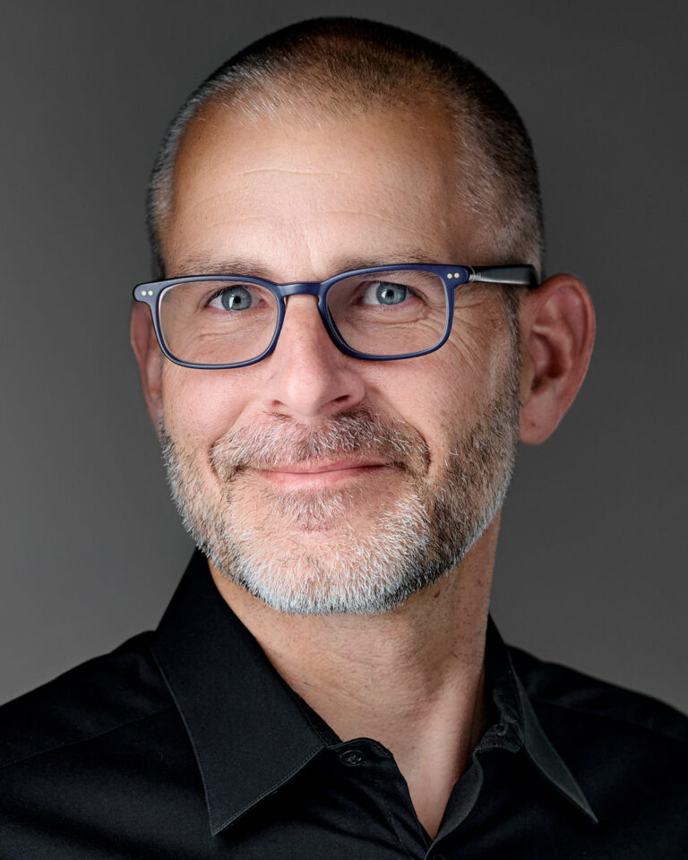 Close-up professional headshot of a man with a salt-and-pepper beard, wearing glasses and a black shirt, photographed in a studio setting in the Bay Area.