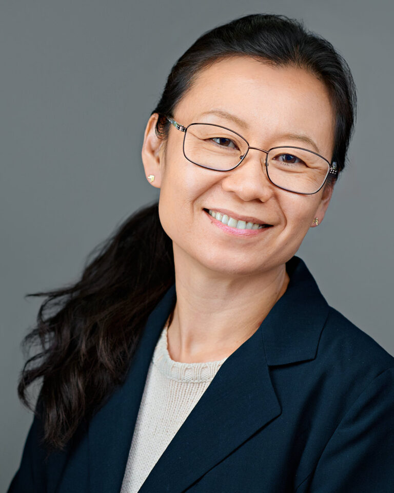 Professional headshot of a woman with long dark hair tied back, wearing glasses and a navy blazer, smiling confidently in a studio setting in Stanford.