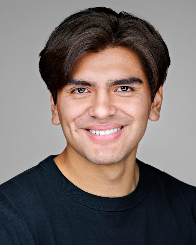 Close-up headshot of a young man with neatly styled dark brown hair, wearing a black shirt and smiling confidently against a neutral gray background in Stanford.