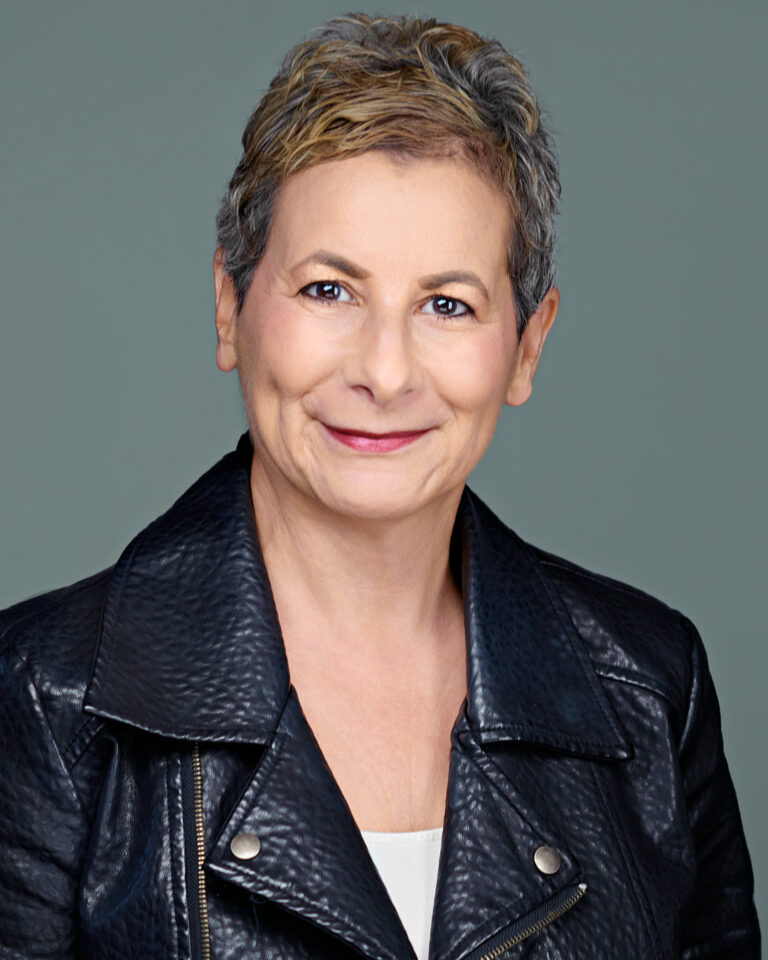 Close-up professional headshot of a mature woman with short, light brown and gray hair, wearing a black leather jacket over a white top, smiling warmly against a soft green background. Photographed in Stanford by Dean Birinyi Photography.