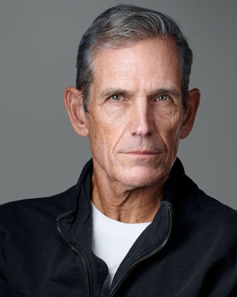 Professional headshot of a mature man with gray hair, wearing a black zip-up jacket over a white t-shirt, against a neutral gray background. Photographed in Stanford by Dean Birinyi Photography.