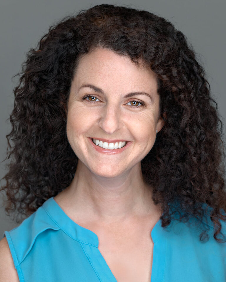 Professional headshot of a smiling woman with curly hair, wearing a blue blouse against a neutral grey background.