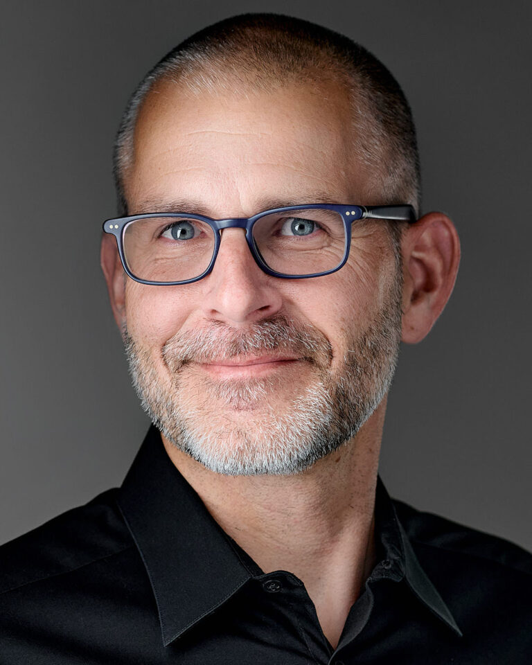 Professional headshot of a man with glasses and a beard, wearing a black shirt, smiling confidently against a gray background in Palo Alto studio.