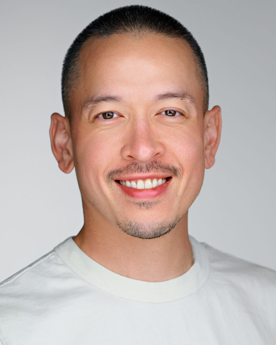 Dean Birinyi, a smiling man with short hair and a goatee, wearing a light-colored t-shirt against a neutral gray background.