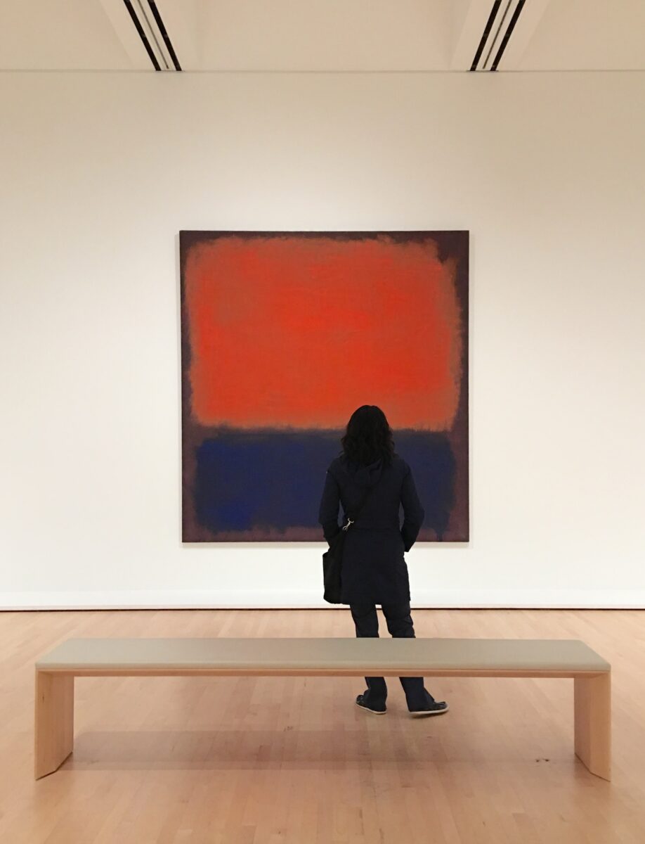 A person dressed in dark clothing stands in front of a large Mark Rothko painting at SFMOMA, taken in 2017. The artwork features a bold red-orange rectangle over a deep blue field, framed by soft, blended edges. A simple wooden bench sits in the foreground on a polished wood floor, emphasizing the quiet, contemplative atmosphere of the gallery space.