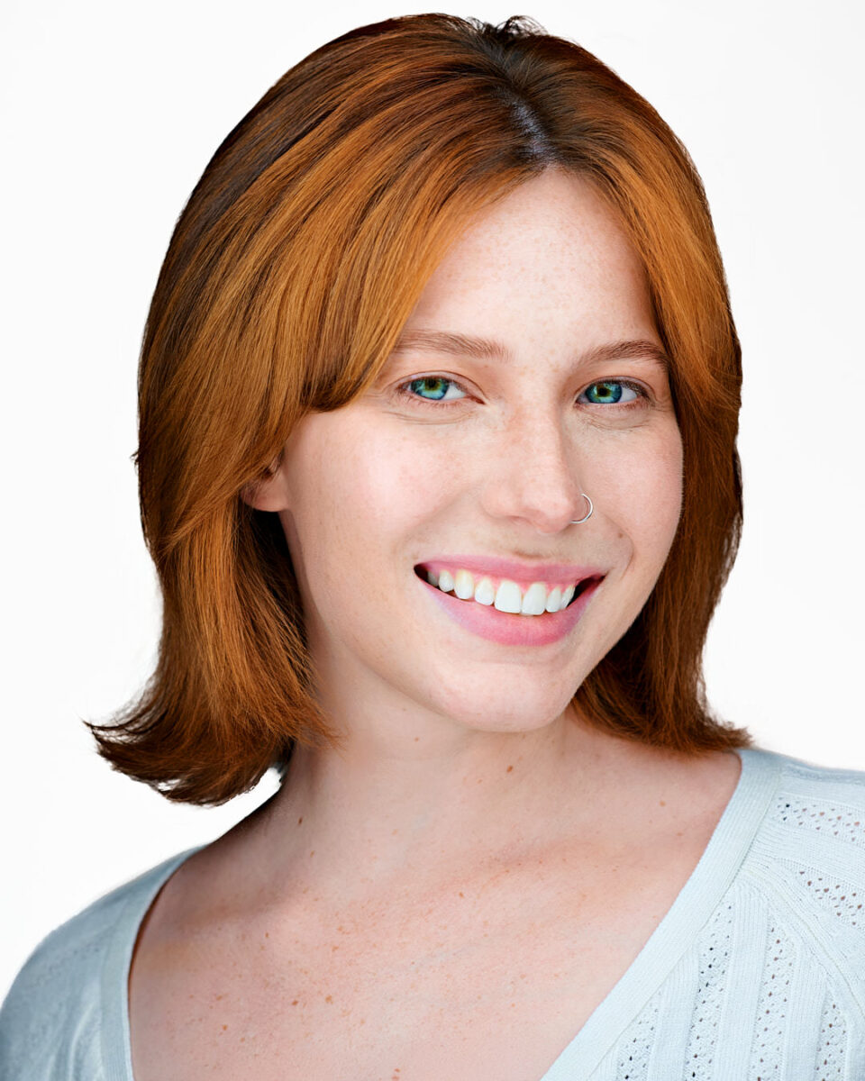 Professional headshot of a confident young woman with red hair, green eyes, and a nose ring, smiling naturally against a clean white background—ideal for authentic, approachable corporate profiles.