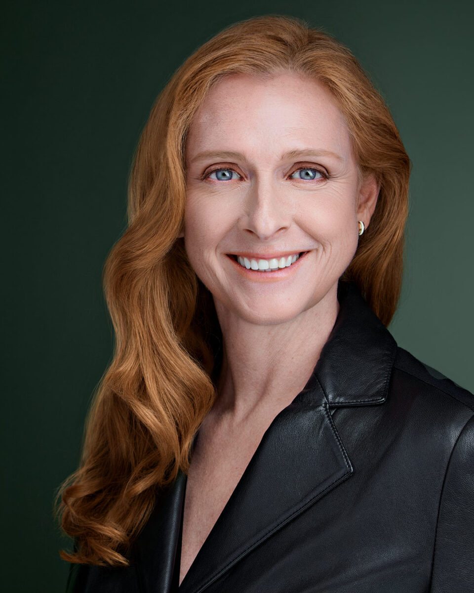 Professional headshot of a red-haired woman in a black leather jacket, smiling confidently against a deep green background. Expertly retouched for a polished yet natural look. Captured by Dean Birinyi in Palo Alto, Silicon Valley.