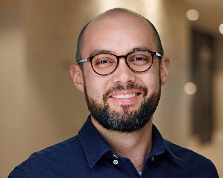 Headshot of a smiling male professional with glasses and a trimmed beard, wearing a navy shirt in an office setting.