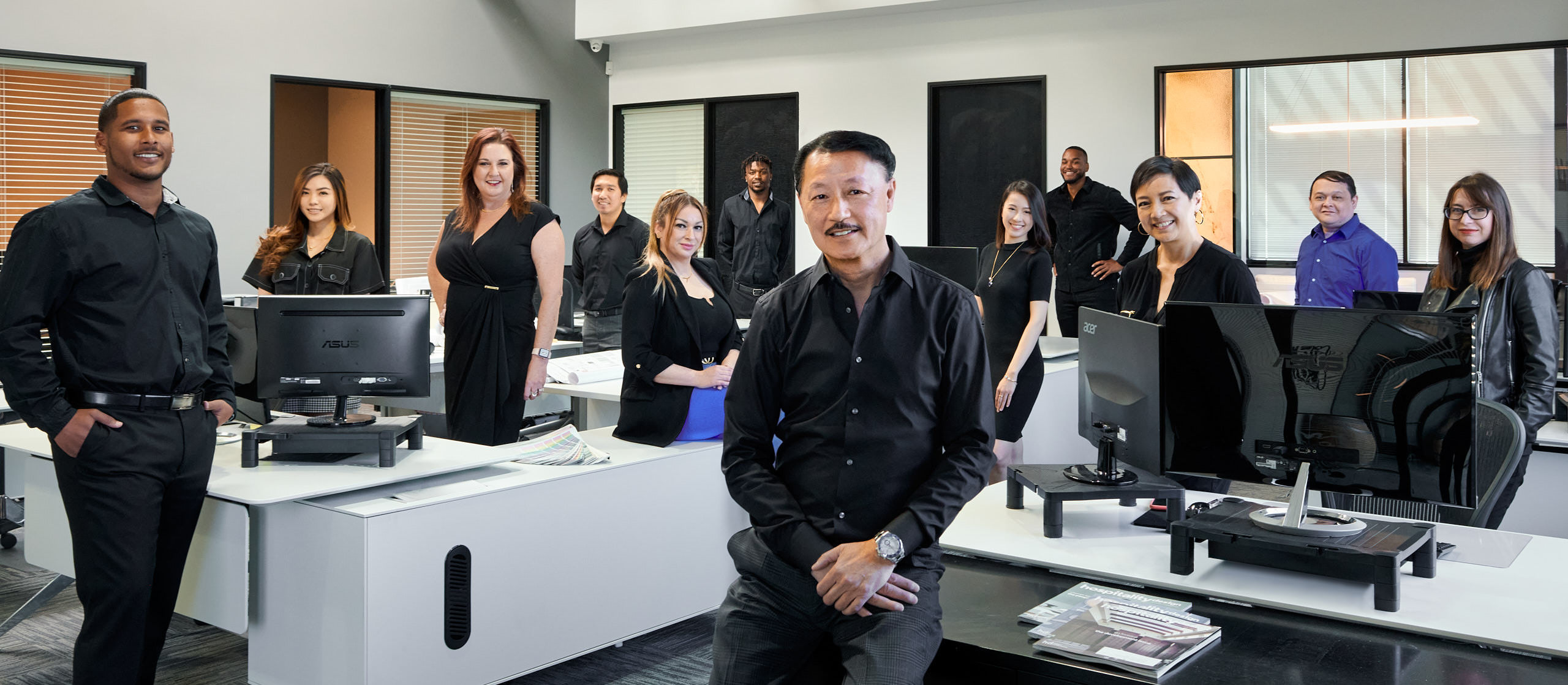 Wide group photo of a diverse team posed confidently throughout a modern office space, dressed in professional black.