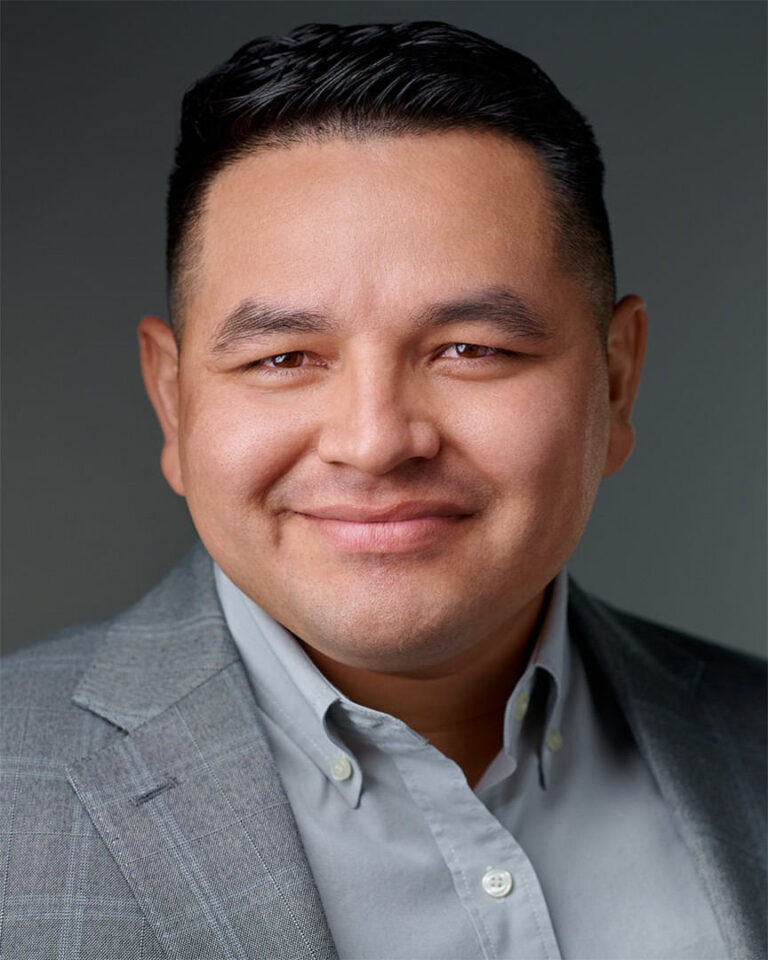 Headshot of a male professional in a gray blazer, smiling confidently against a neutral gray background.