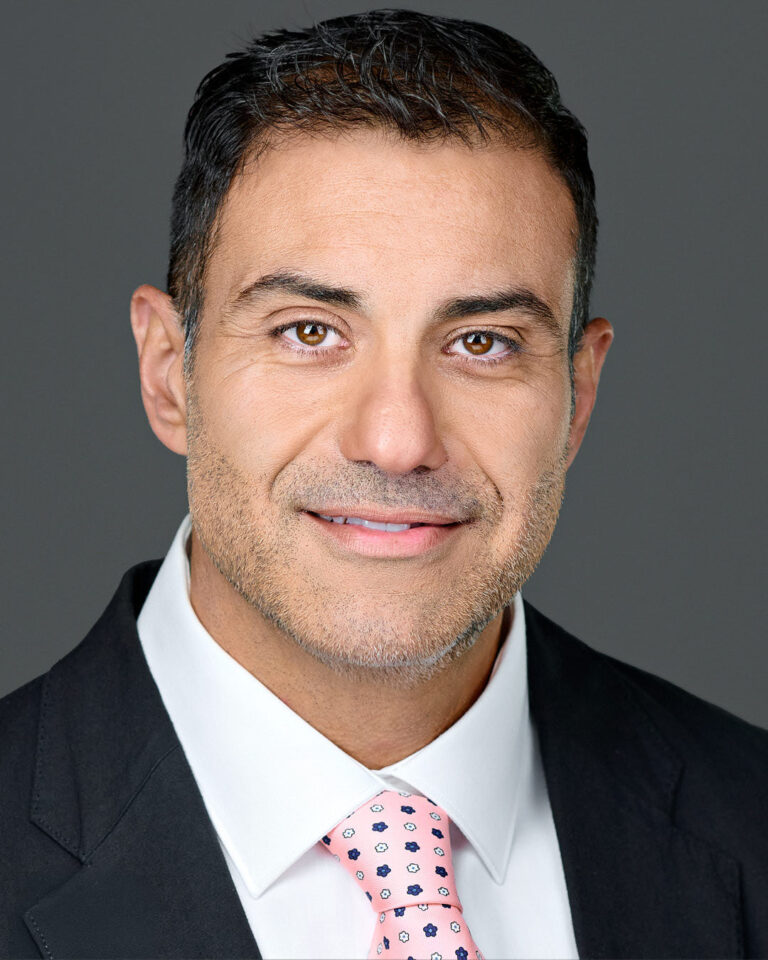 Close-up headshot of a male executive in a suit and pink patterned tie, smiling confidently against a gray background.
