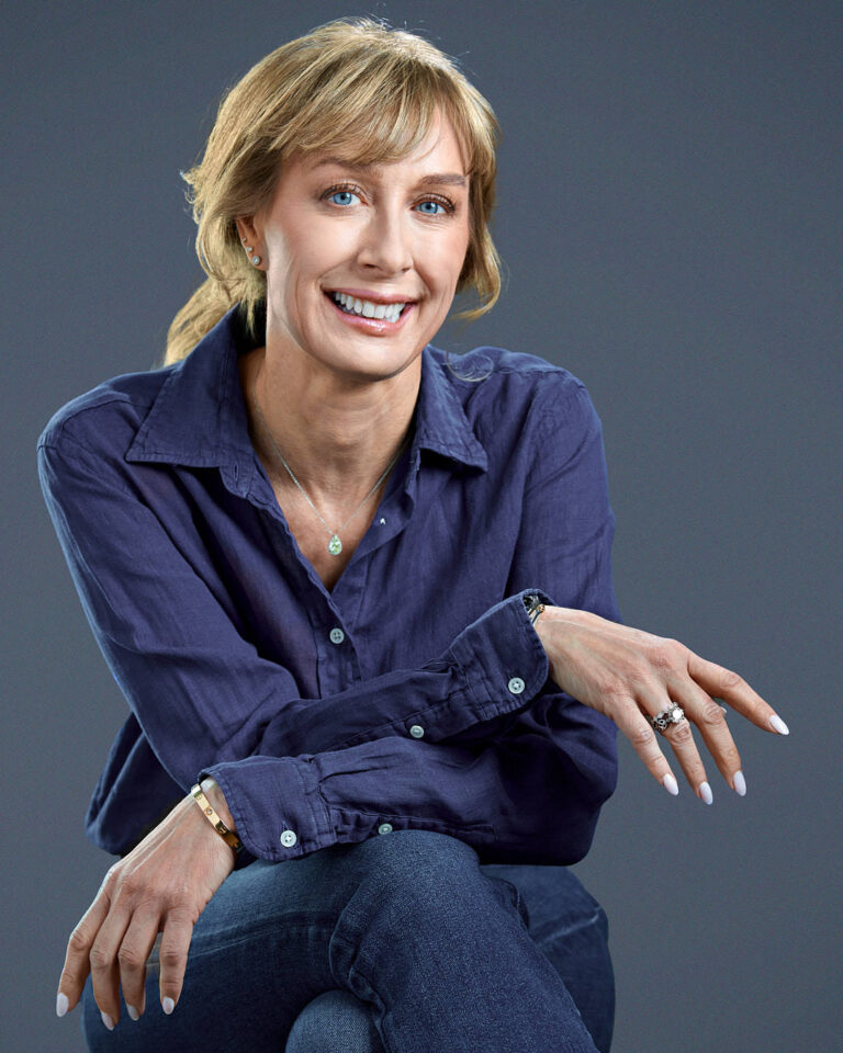 Seated portrait of a woman in a navy blouse and jeans, smiling confidently with arms resting on knee, against a textured gray background.