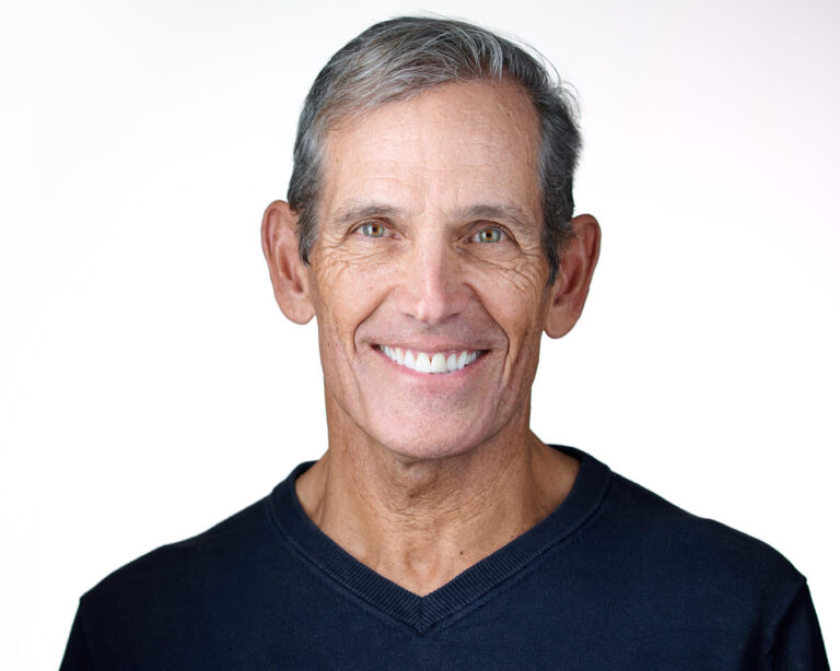 Professional headshot of an older man in a navy sweater, smiling warmly against a white background, projecting confidence and approachability.