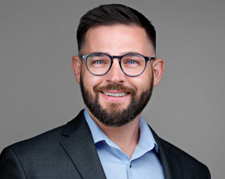 Professional man with dark hair, beard, and glasses wearing a blue shirt and charcoal blazer, smiling confidently in a studio headshot.