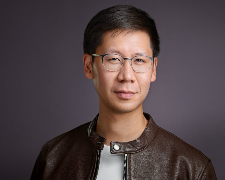 Professional headshot of a man in glasses and a brown leather jacket, photographed against a muted plum background with a composed expression.