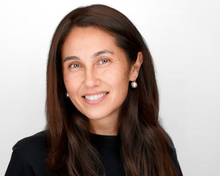 Professional headshot of a woman with long dark hair and pearl earrings, smiling gently against a white background.