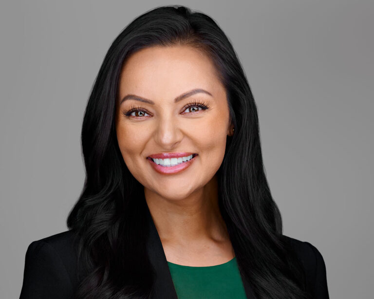 Professional woman with long dark hair in a black blazer and green top, smiling confidently in a studio headshot with a gray background.