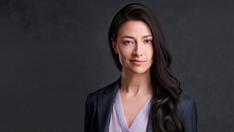 Professional woman with long dark hair wearing a slate blazer and lavender blouse, photographed against a dark gray backdrop with a confident, composed expression.