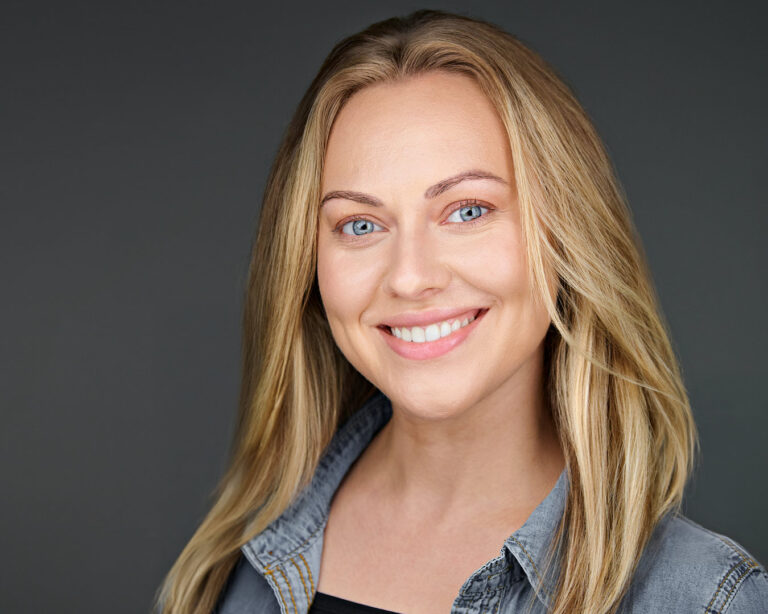 Smiling woman with long blonde hair wearing a denim shirt, photographed against a deep gray studio background with a natural, relaxed expression.